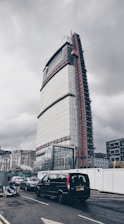 A tall, modern building under construction, partially covered with scaffolding and protective sheeting, dominates the urban scene. Below, several vehicles are waiting at a traffic junction, including a black van. The overcast sky creates a somber atmosphere, and construction signs and barriers are visible along the roadside.