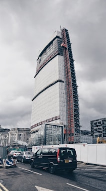 A tall, modern building under construction, partially covered with scaffolding and protective sheeting, dominates the urban scene. Below, several vehicles are waiting at a traffic junction, including a black van. The overcast sky creates a somber atmosphere, and construction signs and barriers are visible along the roadside.