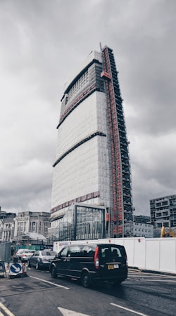 A tall, modern building under construction, partially covered with scaffolding and protective sheeting, dominates the urban scene. Below, several vehicles are waiting at a traffic junction, including a black van. The overcast sky creates a somber atmosphere, and construction signs and barriers are visible along the roadside.