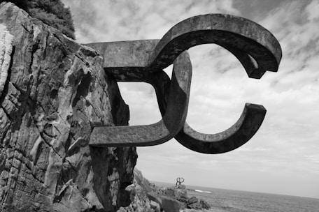 Large metallic sculptures with a curved, abstract design are attached to rocky cliffs by the sea. The sky is mostly cloudy, adding a dramatic backdrop to the scene.