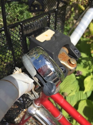 A close-up view of a bicycle handlebar featuring a metallic bell and a black lock. The handlebar is wrapped with white tape, and the bicycle frame is red. The background includes green foliage and a black mesh basket.
