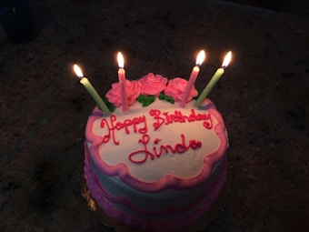 A birthday cake with pink icing decorations and four lit candles on top. The cake has white frosting with pink piping along the edges and roses made of icing. The words 'Happy Birthday Linda' are written in red on the cake.