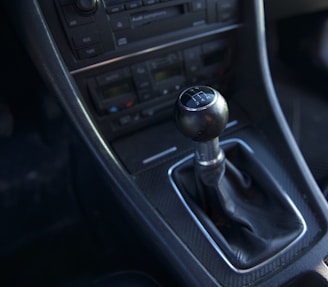 A close-up view of a car's manual gear shift located in the center console. The gear knob is black with silver detailing showing gear positions, surrounded by a leather boot. Below the gear shift are buttons and dials for the car's audio and climate control systems.
