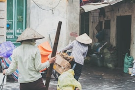 Two people are wearing traditional conical hats in a narrow alleyway, surrounded by bags and a door with Vietnamese text. One person is carrying a large wooden pole, and the other is handling a cart loaded with boxes.