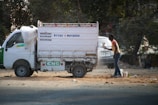 A small white delivery truck with a CNG sticker is parked on a road. The side of the truck has text indicating contact numbers and regions 'Delhi & Haryana'. A person is standing next to the truck on a dusty road, holding a paint sprayer and apparently working on the vehicle. There are trees in the background and another vehicle partially visible.