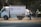 A small white delivery truck with a CNG sticker is parked on a road. The side of the truck has text indicating contact numbers and regions 'Delhi & Haryana'. A person is standing next to the truck on a dusty road, holding a paint sprayer and apparently working on the vehicle. There are trees in the background and another vehicle partially visible.
