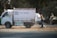 A small white delivery truck with a CNG sticker is parked on a road. The side of the truck has text indicating contact numbers and regions 'Delhi & Haryana'. A person is standing next to the truck on a dusty road, holding a paint sprayer and apparently working on the vehicle. There are trees in the background and another vehicle partially visible.