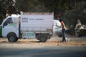 A small white delivery truck with a CNG sticker is parked on a road. The side of the truck has text indicating contact numbers and regions 'Delhi & Haryana'. A person is standing next to the truck on a dusty road, holding a paint sprayer and apparently working on the vehicle. There are trees in the background and another vehicle partially visible.