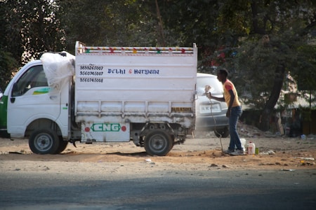A small white delivery truck with a CNG sticker is parked on a road. The side of the truck has text indicating contact numbers and regions 'Delhi & Haryana'. A person is standing next to the truck on a dusty road, holding a paint sprayer and apparently working on the vehicle. There are trees in the background and another vehicle partially visible.