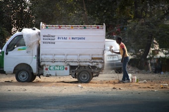 A small white delivery truck with a CNG sticker is parked on a road. The side of the truck has text indicating contact numbers and regions 'Delhi & Haryana'. A person is standing next to the truck on a dusty road, holding a paint sprayer and apparently working on the vehicle. There are trees in the background and another vehicle partially visible.