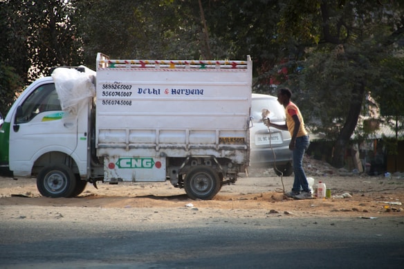 A small white delivery truck with a CNG sticker is parked on a road. The side of the truck has text indicating contact numbers and regions 'Delhi & Haryana'. A person is standing next to the truck on a dusty road, holding a paint sprayer and apparently working on the vehicle. There are trees in the background and another vehicle partially visible.