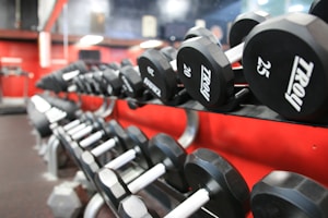 A row of black dumbbells with visible weight labels is neatly arranged on a rack against a red wall in a gym setting. The background includes some blurred gym equipment and mirrors, while the lighting is bright.