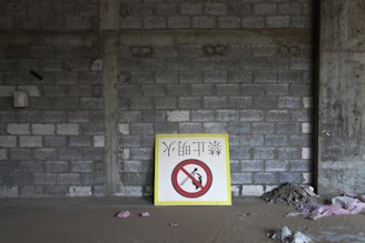 A partially constructed building with concrete block walls and a floor covered in sand and debris. A large sign with bold characters and a warning symbol is propped against the wall.