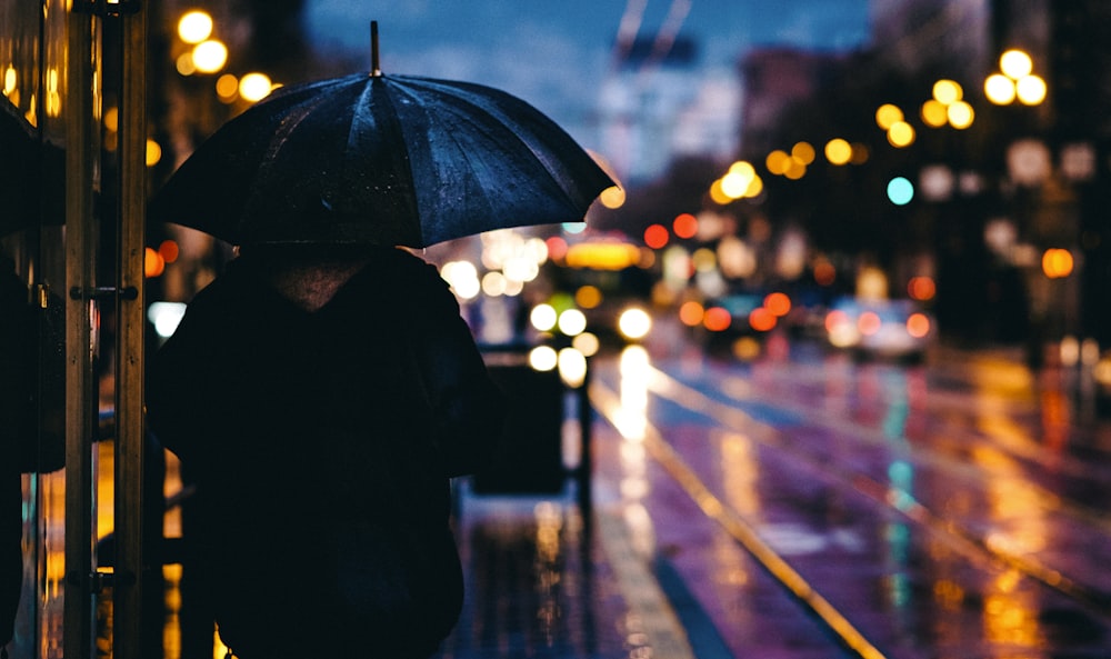 夜間に道路上の車の近くに黒い傘を持って通りを歩いている人