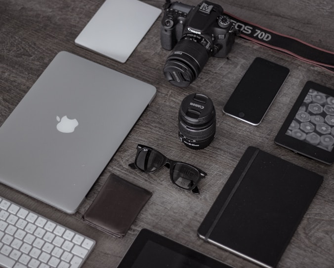 A collection of electronic gadgets and accessories arranged neatly on a wooden surface. Items include a laptop with the Apple logo, a DSLR camera, camera lenses, a smartphone, a wireless keyboard, sunglasses, a leather wallet, a closed notebook, and a tablet device.