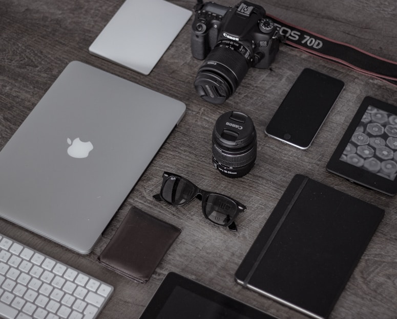 A collection of electronic gadgets and accessories arranged neatly on a wooden surface. Items include a laptop with the Apple logo, a DSLR camera, camera lenses, a smartphone, a wireless keyboard, sunglasses, a leather wallet, a closed notebook, and a tablet device.