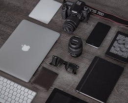 A collection of electronic gadgets and accessories arranged neatly on a wooden surface. Items include a laptop with the Apple logo, a DSLR camera, camera lenses, a smartphone, a wireless keyboard, sunglasses, a leather wallet, a closed notebook, and a tablet device.