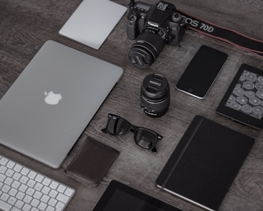 A collection of electronic gadgets and accessories arranged neatly on a wooden surface. Items include a laptop with the Apple logo, a DSLR camera, camera lenses, a smartphone, a wireless keyboard, sunglasses, a leather wallet, a closed notebook, and a tablet device.