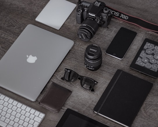 A collection of electronic gadgets and accessories arranged neatly on a wooden surface. Items include a laptop with the Apple logo, a DSLR camera, camera lenses, a smartphone, a wireless keyboard, sunglasses, a leather wallet, a closed notebook, and a tablet device.