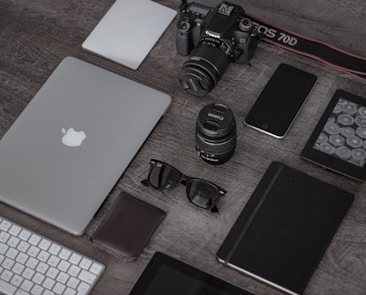 A collection of electronic gadgets and accessories arranged neatly on a wooden surface. Items include a laptop with the Apple logo, a DSLR camera, camera lenses, a smartphone, a wireless keyboard, sunglasses, a leather wallet, a closed notebook, and a tablet device.