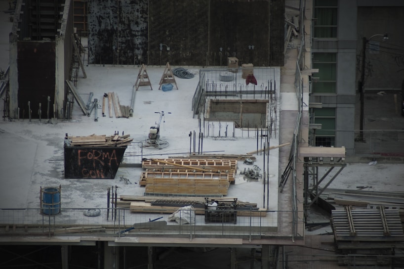 A construction site with scattered materials and tools on a concrete surface. There are wooden planks, metal rods, and a wheelbarrow. Scaffolding surrounds the area. A partially built structure with exposed rebar and openings is evident, alongside a container with graffiti.