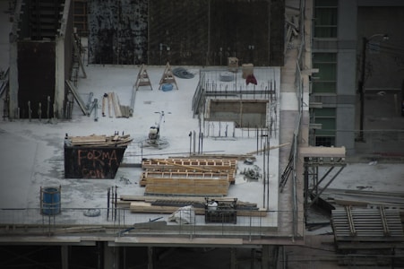 A construction site with scattered materials and tools on a concrete surface. There are wooden planks, metal rods, and a wheelbarrow. Scaffolding surrounds the area. A partially built structure with exposed rebar and openings is evident, alongside a container with graffiti.