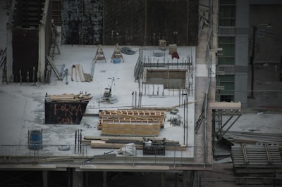 A construction site with scattered materials and tools on a concrete surface. There are wooden planks, metal rods, and a wheelbarrow. Scaffolding surrounds the area. A partially built structure with exposed rebar and openings is evident, alongside a container with graffiti.