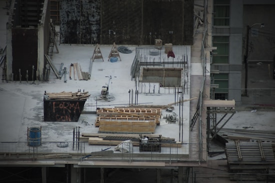 A construction site with scattered materials and tools on a concrete surface. There are wooden planks, metal rods, and a wheelbarrow. Scaffolding surrounds the area. A partially built structure with exposed rebar and openings is evident, alongside a container with graffiti.