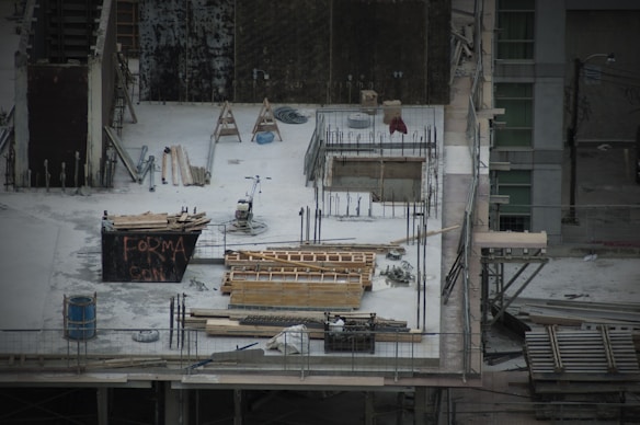 A construction site with scattered materials and tools on a concrete surface. There are wooden planks, metal rods, and a wheelbarrow. Scaffolding surrounds the area. A partially built structure with exposed rebar and openings is evident, alongside a container with graffiti.