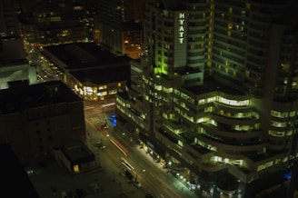 A nighttime cityscape with a view of a high-rise hotel building prominently featuring the name 'Hyatt' in illuminated letters. Surrounding buildings are lit with various windows glowing, and some streetlights are visible. Cars are in motion on the streets below, creating light trails.