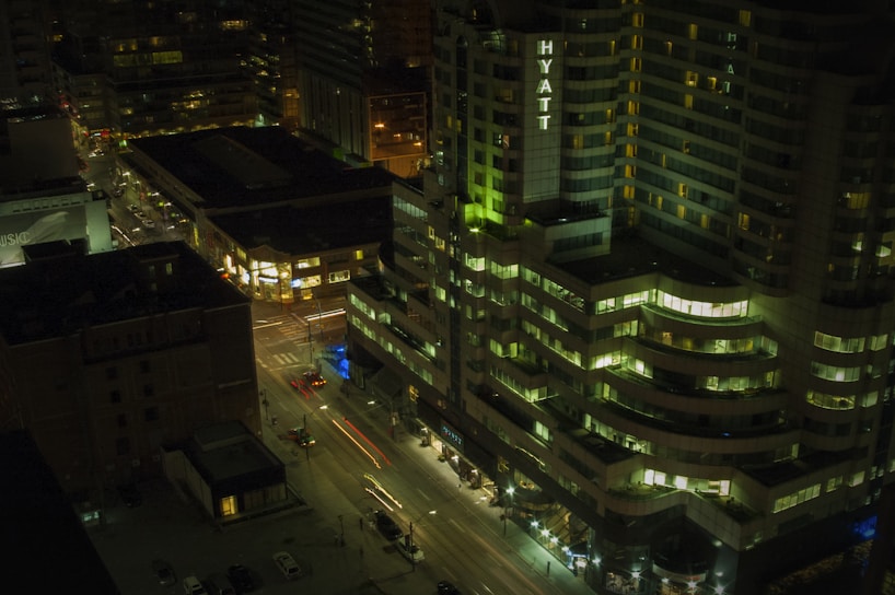 A nighttime cityscape with a view of a high-rise hotel building prominently featuring the name 'Hyatt' in illuminated letters. Surrounding buildings are lit with various windows glowing, and some streetlights are visible. Cars are in motion on the streets below, creating light trails.
