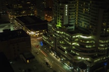 A nighttime cityscape with a view of a high-rise hotel building prominently featuring the name 'Hyatt' in illuminated letters. Surrounding buildings are lit with various windows glowing, and some streetlights are visible. Cars are in motion on the streets below, creating light trails.