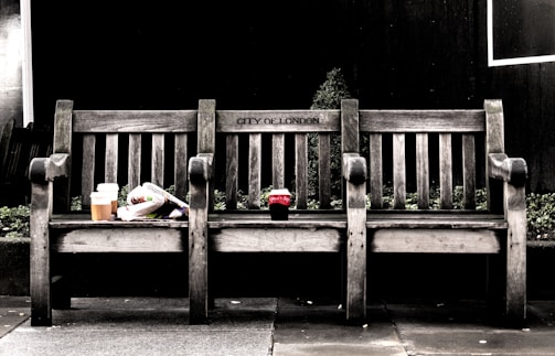 A wooden bench labeled 'City of London' stands in the foreground, with a slightly weathered appearance. On the left side of the bench, there is a disposable coffee cup and a rolled-up newspaper. In the center, a soda cup is placed. The background features dark foliage, adding contrast to the simplicity of the bench.