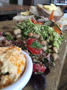 A wooden board is topped with a colorful assortment of food including a quinoa salad with cherry tomatoes, mixed greens, chickpeas, and herbs. A bowl of creamy hummus is visible in the foreground, while slices of bread with butter are stacked in the background.