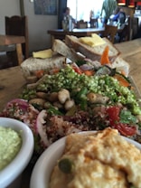 A wooden table set with a variety of fresh food items including a salad filled with green herbs, beans, quinoa, and sliced vegetables. In the foreground, there are bowls containing a creamy green dip and a light-colored hummus. Behind the salad, slices of bread topped with butter are visible. The setting appears to be a cozy cafe with wooden chairs and tables, and a few people can be seen in the background enjoying their time.