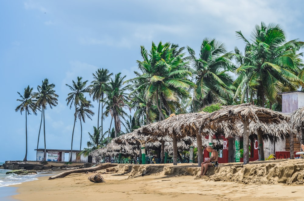 person sitting under brown huts near seashore at daytime