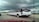 A long, white limousine with tinted windows is driving on a multi-lane highway under a cloudy sky. An American flag is visible on the side along with other vehicles in motion, including a silver SUV. There are signs and a building in the background.