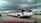 A long, white limousine with tinted windows is driving on a multi-lane highway under a cloudy sky. An American flag is visible on the side along with other vehicles in motion, including a silver SUV. There are signs and a building in the background.