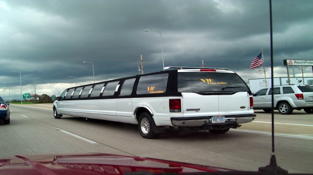A long, white limousine with tinted windows is driving on a multi-lane highway under a cloudy sky. An American flag is visible on the side along with other vehicles in motion, including a silver SUV. There are signs and a building in the background.