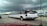 A long, white limousine with tinted windows is driving on a multi-lane highway under a cloudy sky. An American flag is visible on the side along with other vehicles in motion, including a silver SUV. There are signs and a building in the background.