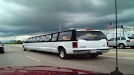 A long, white limousine with tinted windows is driving on a multi-lane highway under a cloudy sky. An American flag is visible on the side along with other vehicles in motion, including a silver SUV. There are signs and a building in the background.