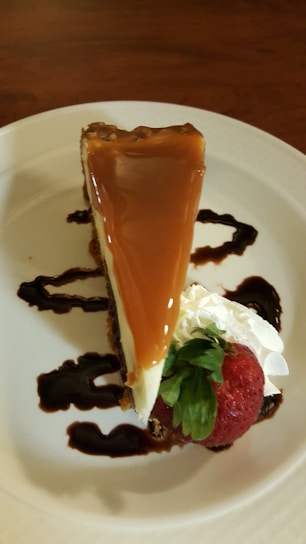 A slice of cheesecake topped with a smooth layer of caramel is elegantly placed on a white plate. It is accented with swirls of chocolate sauce, a dollop of whipped cream, and a fresh strawberry.