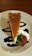 A slice of cheesecake topped with a smooth layer of caramel is elegantly placed on a white plate. It is accented with swirls of chocolate sauce, a dollop of whipped cream, and a fresh strawberry.