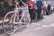 A group of people standing in a line or queue, many carrying backpacks and luggage. The focus is on a vintage-style bicycle leaning against one person's legs. The background shows a cityscape with buildings, suggesting an urban environment.
