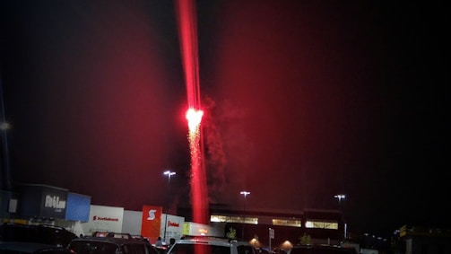 A vertical red flare or firework rises into the night sky, casting a vibrant light. The scene is set in a parking lot with various storefronts visible, including Scotiabank. Several cars are parked in the foreground, and the sky is mostly dark except for the bright red flare.