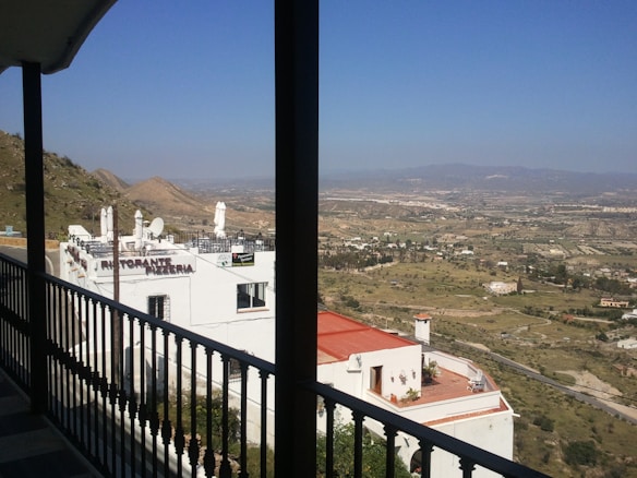 A scenic view of a valley and distant mountains, with a white building labeled 'Ristorante Pizzeria' in the foreground. The landscape is dotted with scattered buildings and greenery, under a clear blue sky.