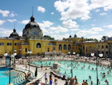 An outdoor public pool filled with people enjoying the water under a sunny sky. The pool is situated in front of a grand, historic building with yellow walls and ornate architectural features. Surrounding the pool are various poolside amenities, such as umbrellas and chairs, where people are lounging and walking around.