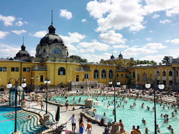 An outdoor public pool filled with people enjoying the water under a sunny sky. The pool is situated in front of a grand, historic building with yellow walls and ornate architectural features. Surrounding the pool are various poolside amenities, such as umbrellas and chairs, where people are lounging and walking around.
