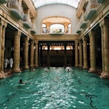 An elegant indoor swimming pool with ornate columns and a high arched glass ceiling. Several people are swimming in the pool, and a few are standing by the edge dressed in white robes. The architectural style is classical with intricate sculptures on the columns. Natural light streams through the skylight, creating reflections on the water.