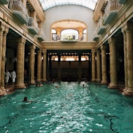 An elegant indoor swimming pool with ornate columns and a high arched glass ceiling. Several people are swimming in the pool, and a few are standing by the edge dressed in white robes. The architectural style is classical with intricate sculptures on the columns. Natural light streams through the skylight, creating reflections on the water.